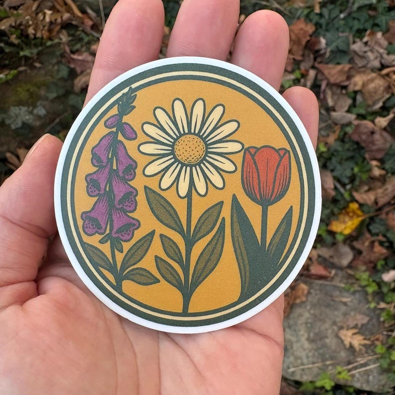 May include: A circular sticker with a floral illustration. The design features a daisy, a tulip, and purple foxgloves set against a yellow background with a white border. The sticker is held in a hand.