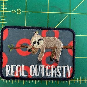 May include: Embroidered patch with a cartoon sloth on a branch. The patch has a navy border, a grey background with red circles, and the words "REAL OUTCASTY" in white.