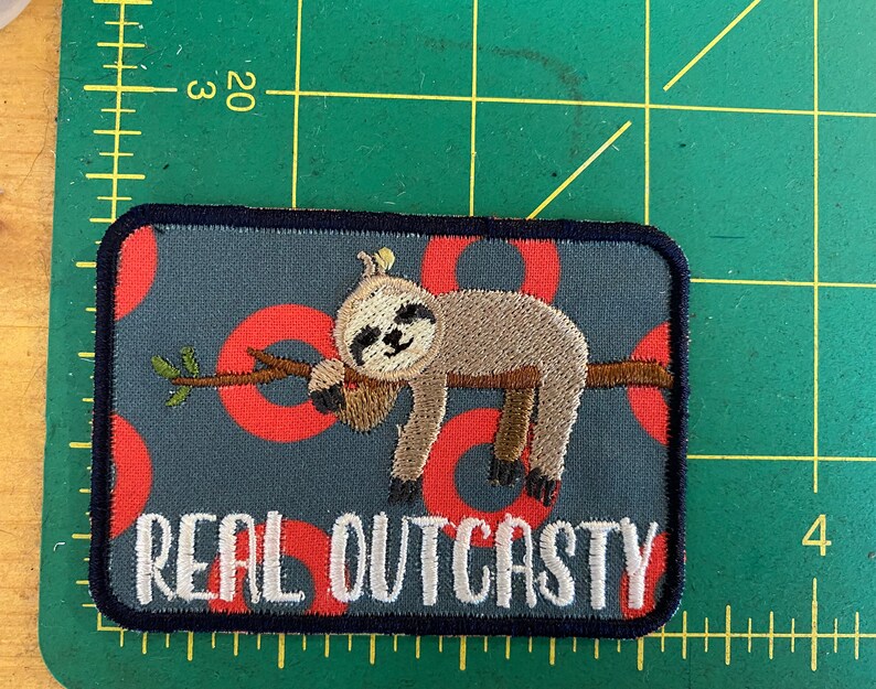 May include: Embroidered patch with a cartoon sloth on a branch. The patch has a navy border, a grey background with red circles, and the words "REAL OUTCASTY" in white.