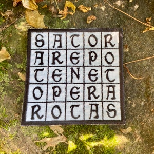 May include: Square patch with the text "SATOR AREPO TENET OPERA ROTAS" in black, block letters. The text is arranged in a grid pattern on a light blue background, with a black border. The patch is likely embroidered or printed.