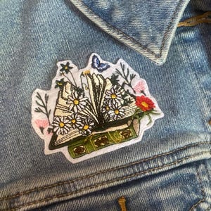 May include: Embroidered patch featuring an open book overflowing with daisies, other flowers, and a butterfly. The patch has a white border and is attached to a denim jacket. The book is green with gold accents.