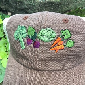 May include: A brown baseball cap with a white mesh back. The front of the cap features embroidered vegetables: broccoli, radishes, cabbage, and carrots. The cap has a curved brim and a button on top.
