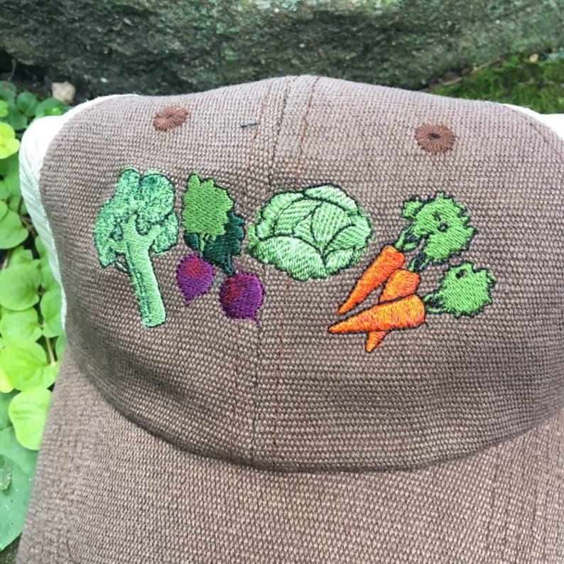 May include: A brown baseball cap with a white mesh back. The front of the cap features embroidered vegetables: broccoli, radishes, cabbage, and carrots. The cap has a curved brim and a button on top.