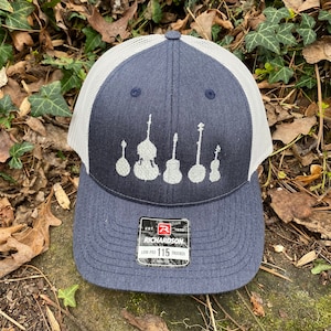 Pode incluir: Bon&eacute; de caminhoneiro azul marinho com parte traseira em malha branca. A frente do bon&eacute; apresenta um design bordado de instrumentos de cordas. O bon&eacute; tem uma aba curva e uma etiqueta que diz "RICHARDSON LOW-PRO 115 TRUCKER".