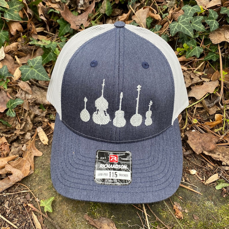 Pode incluir: Bon&eacute; de caminhoneiro azul marinho com parte traseira em malha branca. A frente do bon&eacute; apresenta um design bordado de instrumentos de cordas. O bon&eacute; tem uma aba curva e uma etiqueta que diz "RICHARDSON LOW-PRO 115 TRUCKER".