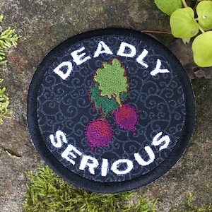 May include: Round black patch with the words "DEADLY SERIOUS" in white, surrounding an embroidered image of beets. The beets are purple and red with green leaves. The background is a dark fabric with a swirling pattern.