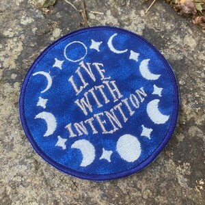 May include: A circular blue patch featuring white embroidered celestial designs, such as moons, stars, and a circle. The words "LIVE WITH INTENTION" are embroidered in white. This patch is likely designed for application on clothing or accessories.