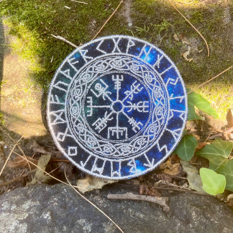 May include: Circular embroidered patch with a blue and white Viking compass design, or Vegv&iacute;sir. The patch has a dark blue, starry background, and white runes around the edge. The design is framed by a white border. The patch is a symbol of guidance.