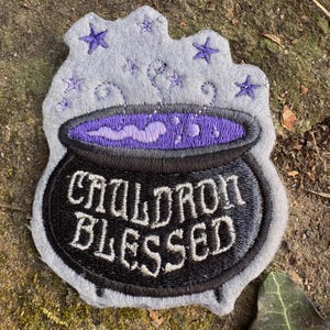 May include: Embroidered patch featuring a black cauldron with the words "CAULDRON BLESSED" in white. The cauldron has a purple liquid and is surrounded by gray steam and purple stars. The patch is on a gray background.