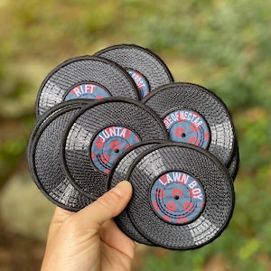May include: A collection of black, circular embroidered patches resembling vinyl records. Each patch features a central label with text in red and white on a blue background. The patches are held in a hand against a blurred green background.