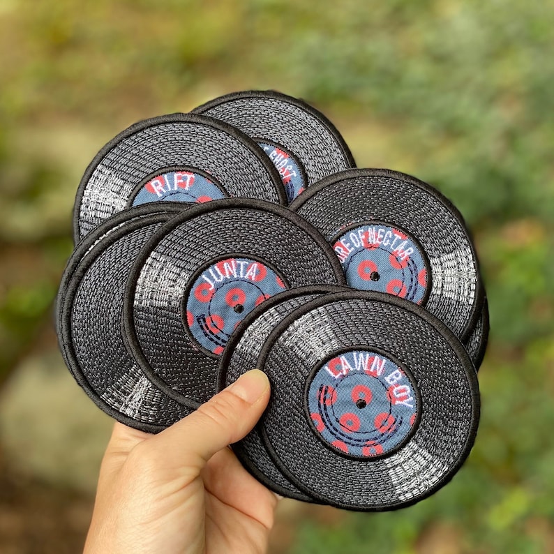 May include: A collection of black, circular embroidered patches resembling vinyl records. Each patch features a central label with text in red and white on a blue background. The patches are held in a hand against a blurred green background.