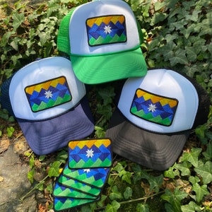 May include: A collection of trucker hats with white fronts, mesh backs, and colorful embroidered patches. The patches feature a geometric design in blue, green, yellow, and a white star. One hat has a green mesh back, another has a navy blue back, and another has a black back.