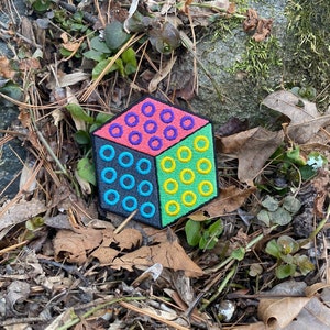 Puede incluir: Un parche bordado colorido en forma de cubo, dividido en cuatro secciones. Cada secci&oacute;n presenta un color y un patr&oacute;n diferente: rosa con c&iacute;rculos morados, verde con c&iacute;rculos amarillos, azul con c&iacute;rculos turquesa y gris con c&iacute;rculos turquesa.