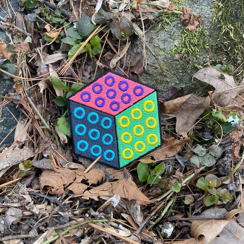 Puede incluir: Un parche bordado colorido en forma de cubo, dividido en cuatro secciones. Cada secci&oacute;n presenta un color y un patr&oacute;n diferente: rosa con c&iacute;rculos morados, verde con c&iacute;rculos amarillos, azul con c&iacute;rculos turquesa y gris con c&iacute;rculos turquesa.