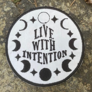 May include: A circular white patch with black embroidery. The patch displays the words "LIVE WITH INTENTION" in a decorative font, encircled by crescent moons, stars, and a full moon. The patch is edged with a black border.