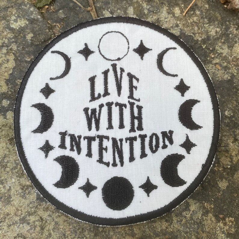 May include: A circular white patch with black embroidery. The patch displays the words "LIVE WITH INTENTION" in a decorative font, encircled by crescent moons, stars, and a full moon. The patch is edged with a black border.