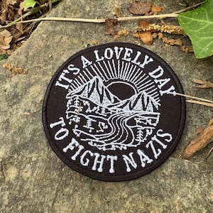 May include: A round, black patch with white embroidered text and a landscape design. The text reads "It's a lovely day to fight Nazis." The landscape features mountains, a river, and a sunburst.