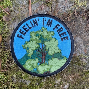 May include: A round patch with the text "FEELIN' I'M FREE" in black lettering on a blue background. The patch features a detailed embroidered tree in shades of green and brown, with a black border.