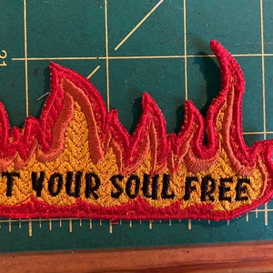 May include: Embroidered flame-shaped patch, featuring red and yellow thread. The text "SET YOUR SOUL FREE" is stitched in black. The patch is displayed on a green cutting mat.