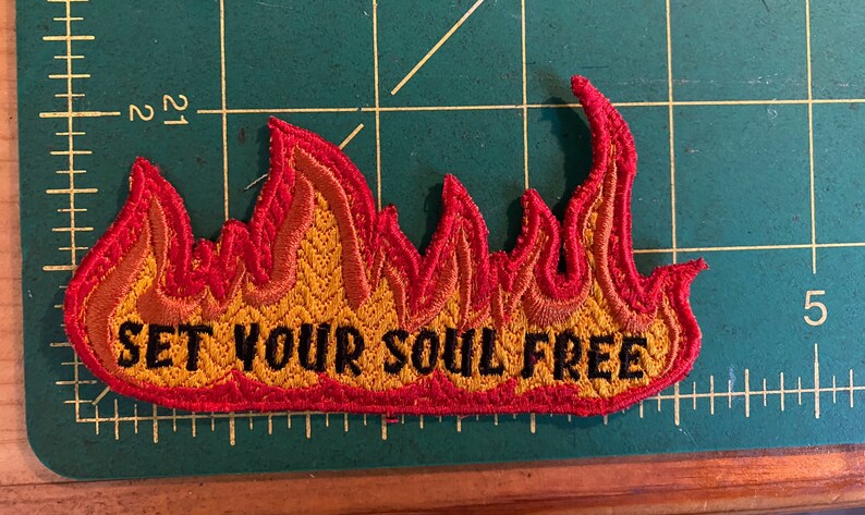 May include: Embroidered flame-shaped patch, featuring red and yellow thread. The text "SET YOUR SOUL FREE" is stitched in black. The patch is displayed on a green cutting mat.