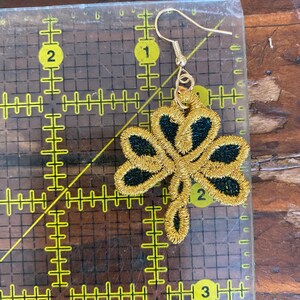 Pode incluir: Um brinco de trevo bordado dourado com detalhes em preto. O brinco tem um gancho dourado. O brinco tem aproximadamente 5 cm de comprimento.