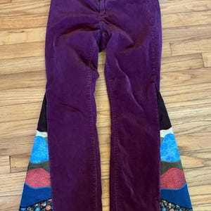 Op de afbeelding: Pruimkleurige corduroy broek met een uitlopend pijp ontwerp. De uitlopende secties hebben een patchwork ontwerp met verschillende kleuren en bloemmotieven, wat een unieke uitstraling cre&euml;ert. De broek ligt plat op een houten oppervlak.
