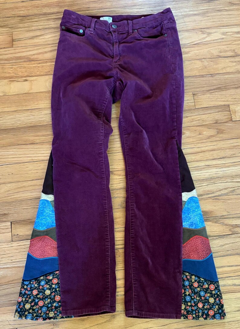 Op de afbeelding: Pruimkleurige corduroy broek met een uitlopend pijp ontwerp. De uitlopende secties hebben een patchwork ontwerp met verschillende kleuren en bloemmotieven, wat een unieke uitstraling cre&euml;ert. De broek ligt plat op een houten oppervlak.