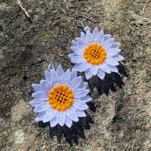 May include: A pair of daisy-shaped earrings. Each earring features a white, lace-like petal design surrounding a yellow center. The earrings hang from silver-colored hooks. The earrings are set against a natural, textured background.