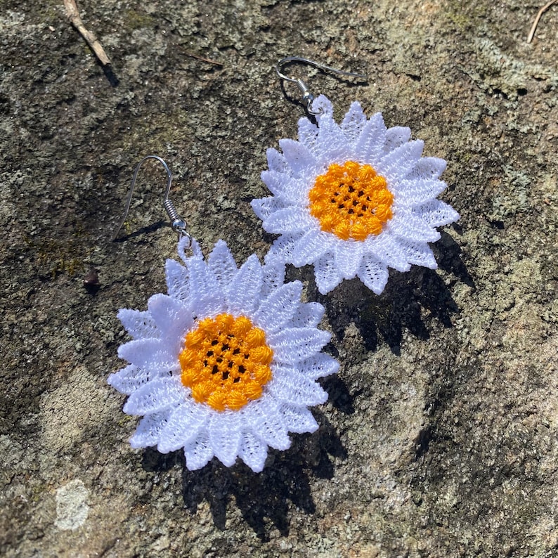 May include: A pair of daisy-shaped earrings. Each earring features a white, lace-like petal design surrounding a yellow center. The earrings hang from silver-colored hooks. The earrings are set against a natural, textured background.