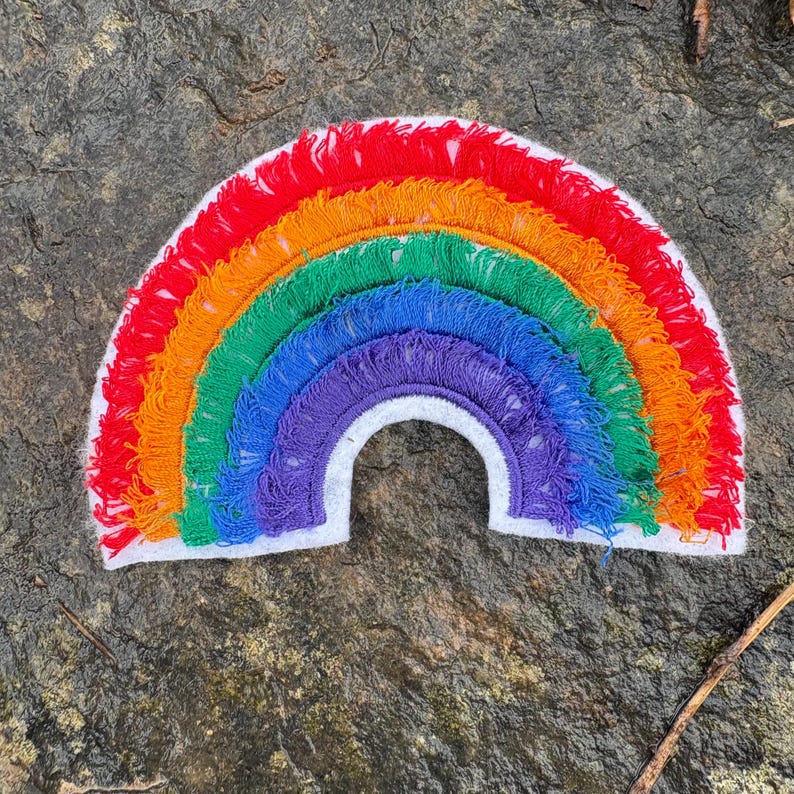 May include: A colorful rainbow-shaped patch with red, orange, yellow, green, blue, and purple fringe. The rainbow is outlined with white fabric. The patch is likely for clothing or accessories.