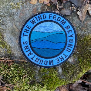 May include: A circular embroidered patch with the text "THE WIND FROM BEYOND THE MOUNTAINS" in blue. The patch depicts a mountain scene in shades of blue and has a black border. It rests on a rock covered in moss.