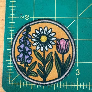 Pu&ograve; includere: Toppa ricamata rotonda con un design floreale. La toppa ha uno sfondo giallo e presenta una margherita, un tulipano e digitali viola, con foglie e steli verdi. La toppa &egrave; bordata con cuciture color crema.