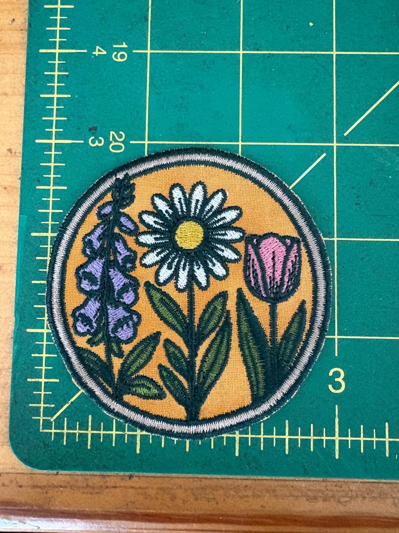 Pu&ograve; includere: Toppa ricamata rotonda con un design floreale. La toppa ha uno sfondo giallo e presenta una margherita, un tulipano e digitali viola, con foglie e steli verdi. La toppa &egrave; bordata con cuciture color crema.