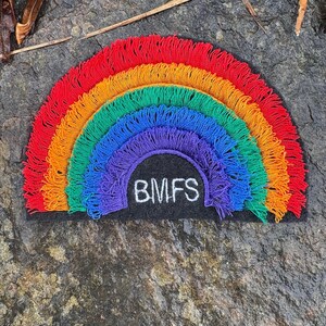 May include: A colorful rainbow patch with red, orange, yellow, green, blue, and purple layers. The patch has a black base with the letters "BMFS" embroidered in white. The rainbow design is made of fringed threads.