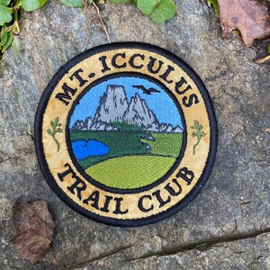 Op de afbeelding: Ronde geborduurde patch met de tekst "MT. ICCULUS TRAIL CLUB". Het ontwerp toont een bergketen, een meer en een groen veld onder een blauwe hemel. De patch heeft een beige rand en ligt op een stenen oppervlak.