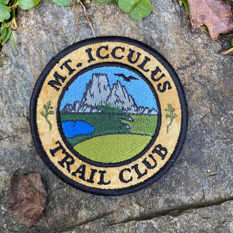 Op de afbeelding: Ronde geborduurde patch met de tekst "MT. ICCULUS TRAIL CLUB". Het ontwerp toont een bergketen, een meer en een groen veld onder een blauwe hemel. De patch heeft een beige rand en ligt op een stenen oppervlak.