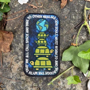 May include: A rectangular patch with a dark blue background and a design featuring a stack of turtles supporting a globe. The patch has text that reads "THAT WE ALL CALL SPACE AND TIME" and "OUR SOULS MUST ROAM TO AND THROUGH THE MYTH."