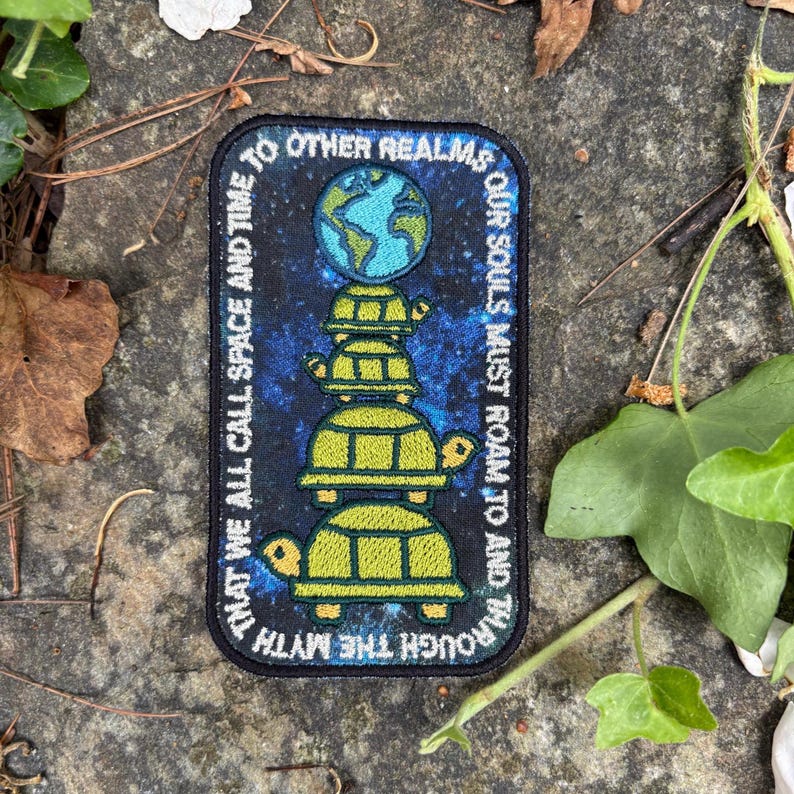 May include: A rectangular patch with a dark blue background and a design featuring a stack of turtles supporting a globe. The patch has text that reads "THAT WE ALL CALL SPACE AND TIME" and "OUR SOULS MUST ROAM TO AND THROUGH THE MYTH."