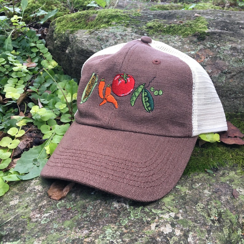 May include: A brown baseball cap with a white mesh back. The front of the cap features embroidered vegetables: corn, chili peppers, a tomato, and peas. The cap is sitting on a rock.