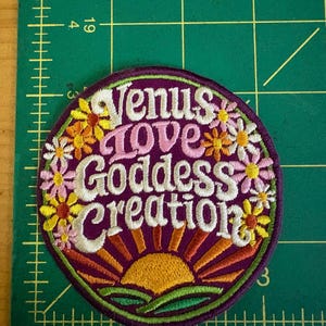 May include: A circular embroidered patch with the text "Venus Love Goddess Creation" in white script. The patch has a purple background, a floral border, and a sunrise design. The flowers are in shades of yellow, orange, and pink.