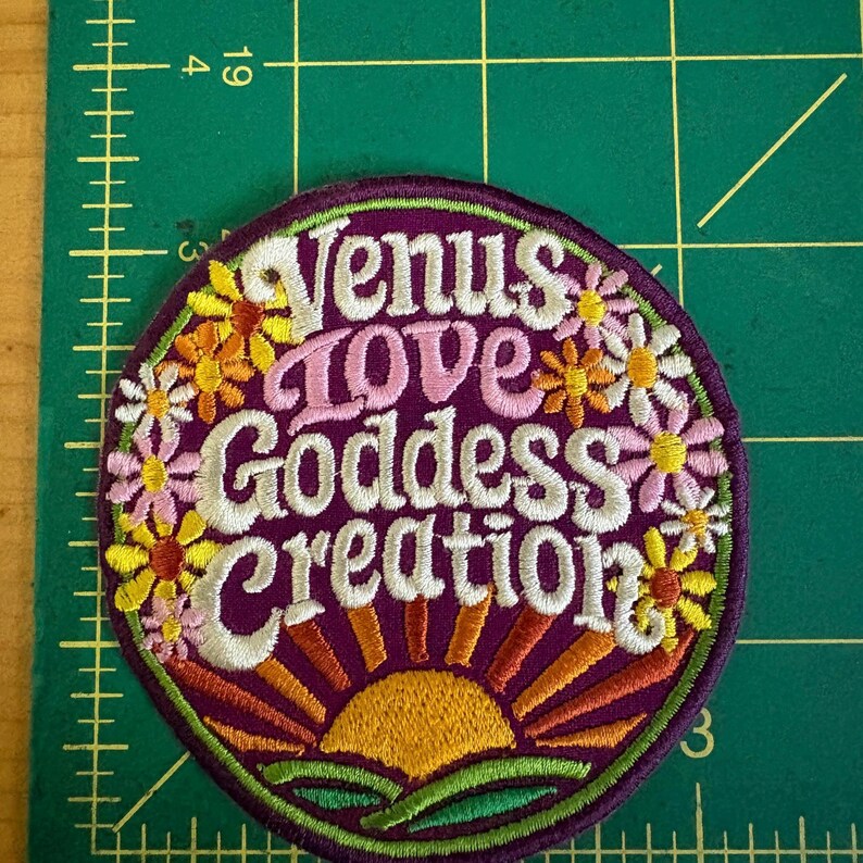 May include: A circular embroidered patch with the text "Venus Love Goddess Creation" in white script. The patch has a purple background, a floral border, and a sunrise design. The flowers are in shades of yellow, orange, and pink.