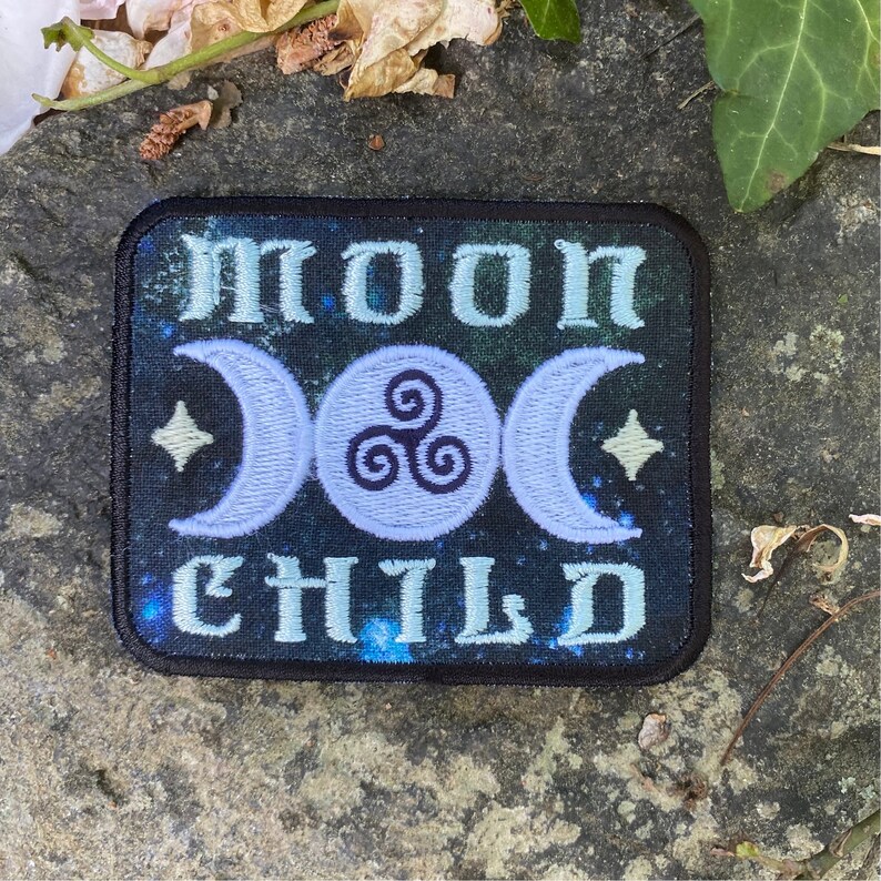 Op de afbeelding: Geborduurde patch met de woorden "MOON CHILD" in lichtblauw, tegen een donkerblauwe en groene sterrenachtergrond. Voorzien van een drievoudig maansymbool en twee sterren. Zwarte rand.