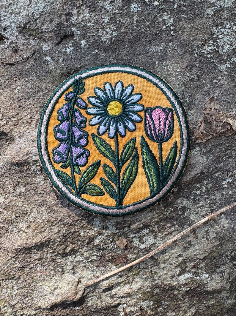 Pu&ograve; includere: Toppa rotonda ricamata con una margherita, un tulipano e fiori di digitale purpurea. La toppa ha uno sfondo giallo e un bordo verde. I fiori sono dettagliati con foglie e steli verdi. La toppa &egrave; rotonda e sembra cucita.