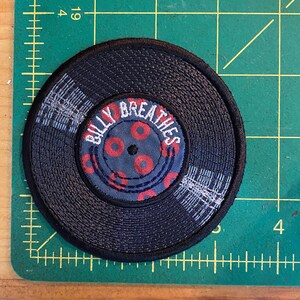 May include: A round, black embroidered patch resembling a vinyl record. The center features the text "BILLY BREATHES" in white, surrounded by red circles on a blue background. The patch has a textured appearance.