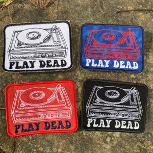 May include: Four rectangular patches featuring a turntable design with the text "PLAY DEAD". The patches are in white, blue, red, and black, with contrasting thread colors for the turntable and text.