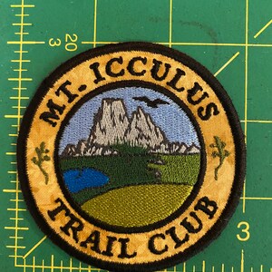 Op de afbeelding: Geborduurde ronde patch met de tekst "MT. ICCULUS TRAIL CLUB". Het ontwerp toont een berglandschap met een blauwe lucht, een meer en groene en beige accenten. Een zwarte rand omringt het ontwerp.
