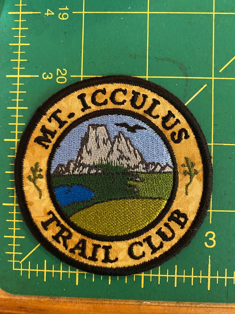 Op de afbeelding: Geborduurde ronde patch met de tekst "MT. ICCULUS TRAIL CLUB". Het ontwerp toont een berglandschap met een blauwe lucht, een meer en groene en beige accenten. Een zwarte rand omringt het ontwerp.