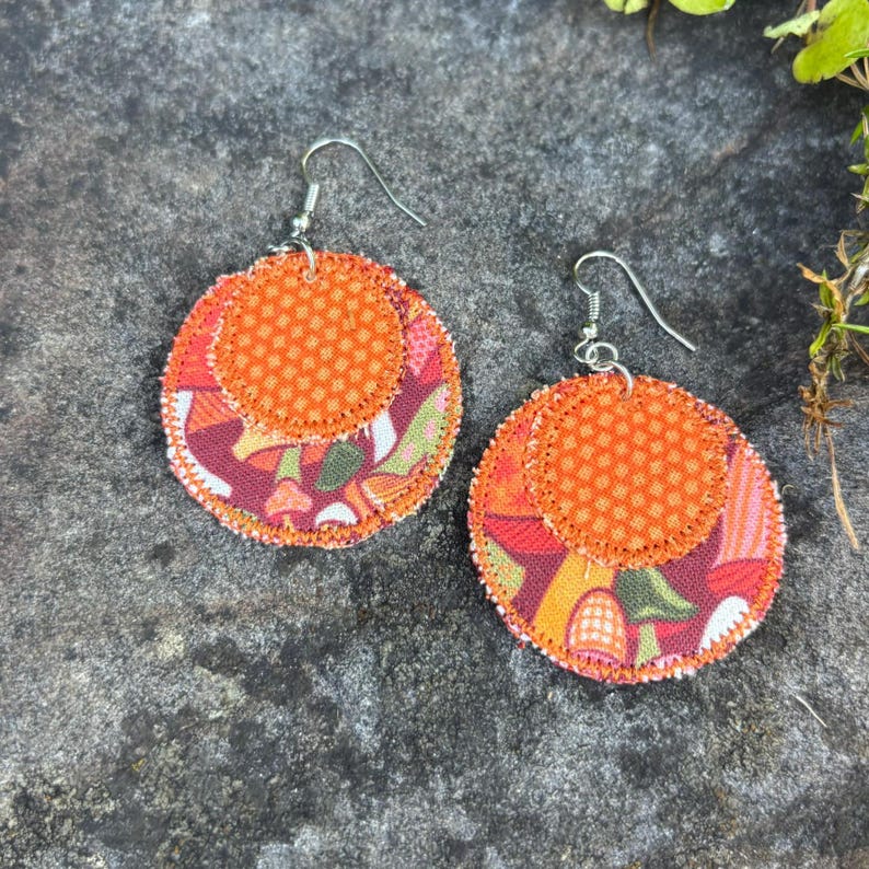 Pu&ograve; includere: Un paio di orecchini in tessuto fatti a mano. Ogni orecchino presenta una base circolare con un colorato motivo floreale nei toni del rosso, arancione e verde. Un cerchio arancione pi&ugrave; piccolo a pois &egrave; cucito sopra, con ganci per orecchie color argento.