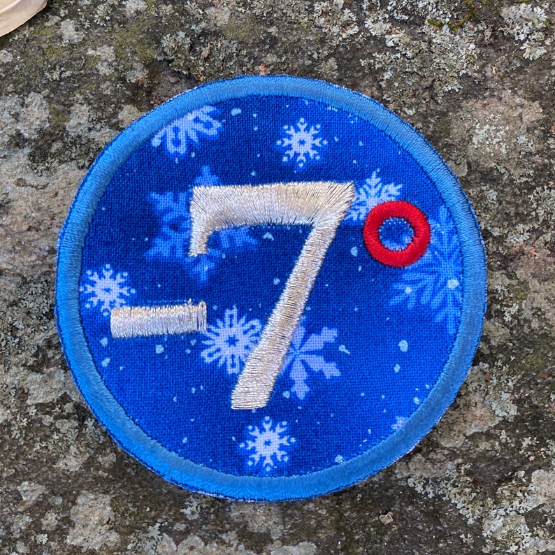 Op de afbeelding: Een ronde, blauwe stoffen patch met een sneeuwvlokpatroon en het geborduurde nummer -7 in zilver. Een kleine rode cirkel staat naast het nummer. De patch heeft een lichtblauwe rand.