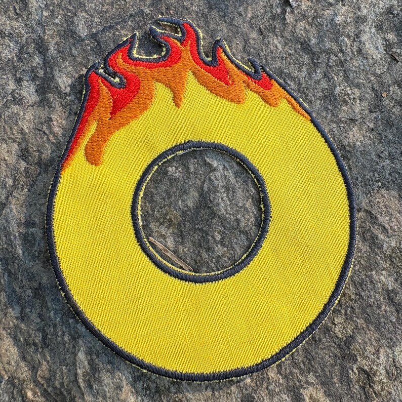 Puede incluir: Parche bordado amarillo con forma de la letra "O" con un dise&ntilde;o de llama. Las llamas son rojas, naranjas y amarillas. El parche tiene un borde azul oscuro y est&aacute; sobre una superficie de piedra gris.